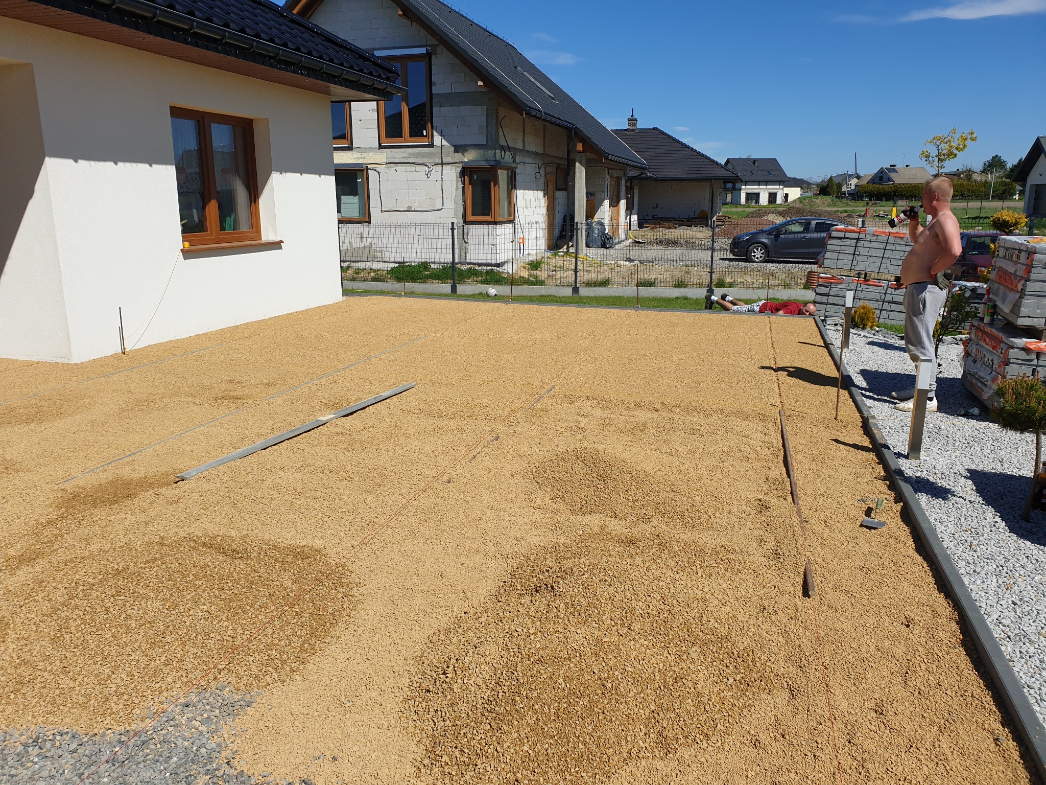 Przygotowanie podłoża pod kostkę brukową: wyrównana warstwa piasku na dużej powierzchni przed domem w trakcie prac, widoczne profile i sznurki wyznaczające obszar układania, w tle budynek w trakcie...