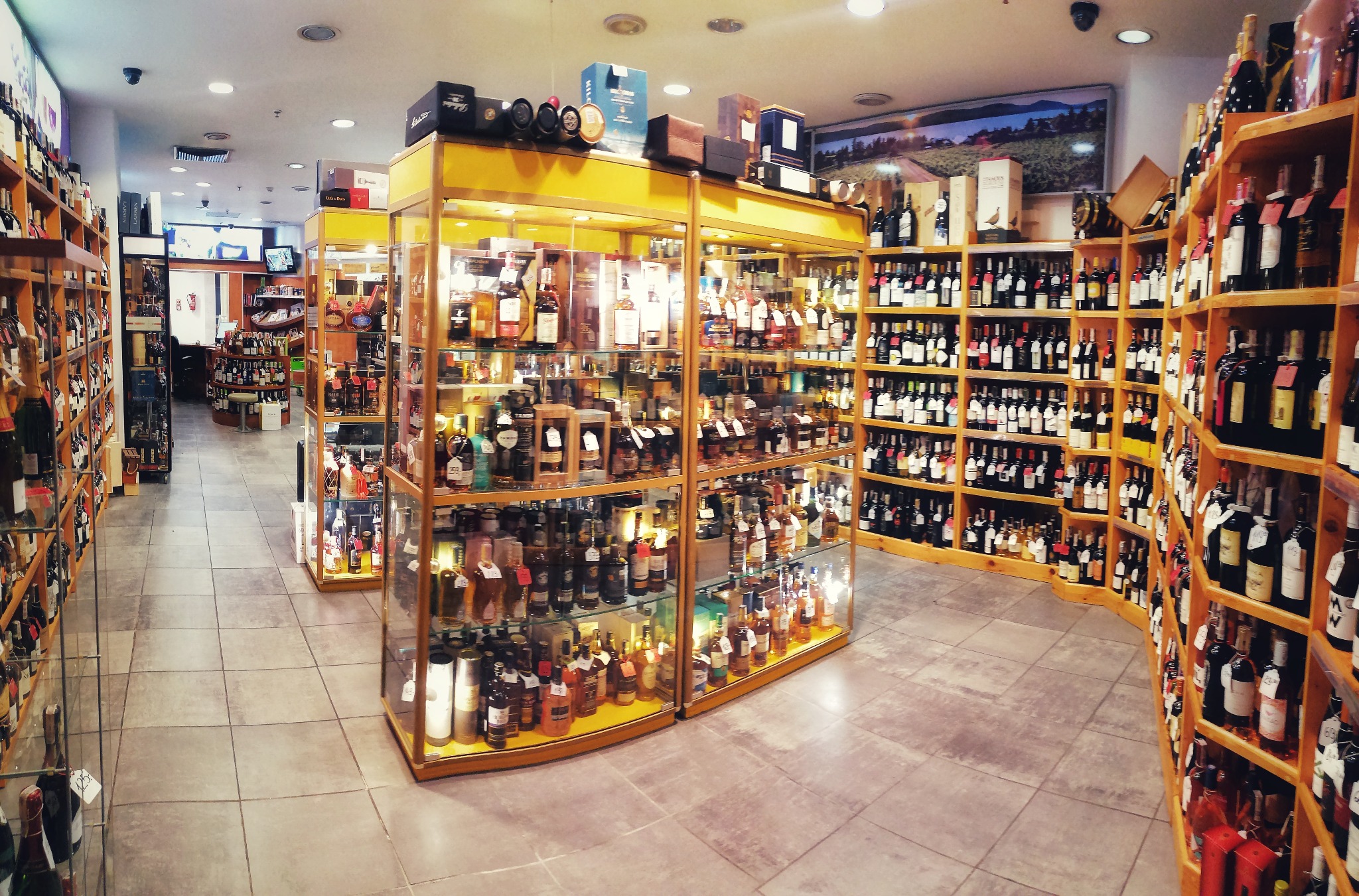 Wnętrze przestronnego sklepu z alkoholami: regały i przeszklone gabloty wypełnione różnorodnymi butelkami i opakowaniami, widok z perspektywy klienta, na podłodze jasne płytki.