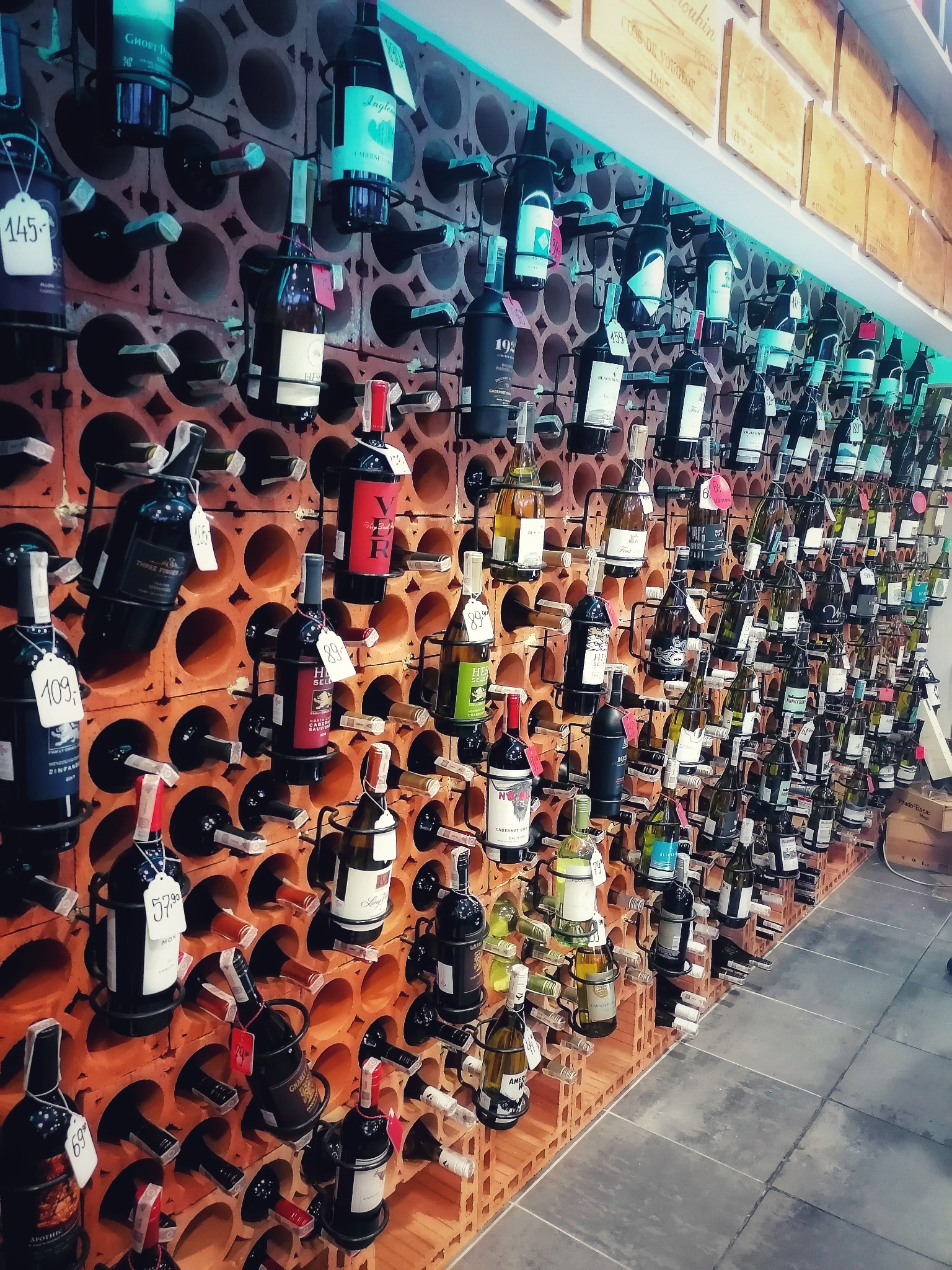 Ściana z terakoty z licznymi butelkami wina w metalowych uchwytach, każda z ceną, w tle drewniane skrzynki i oświetlenie LED