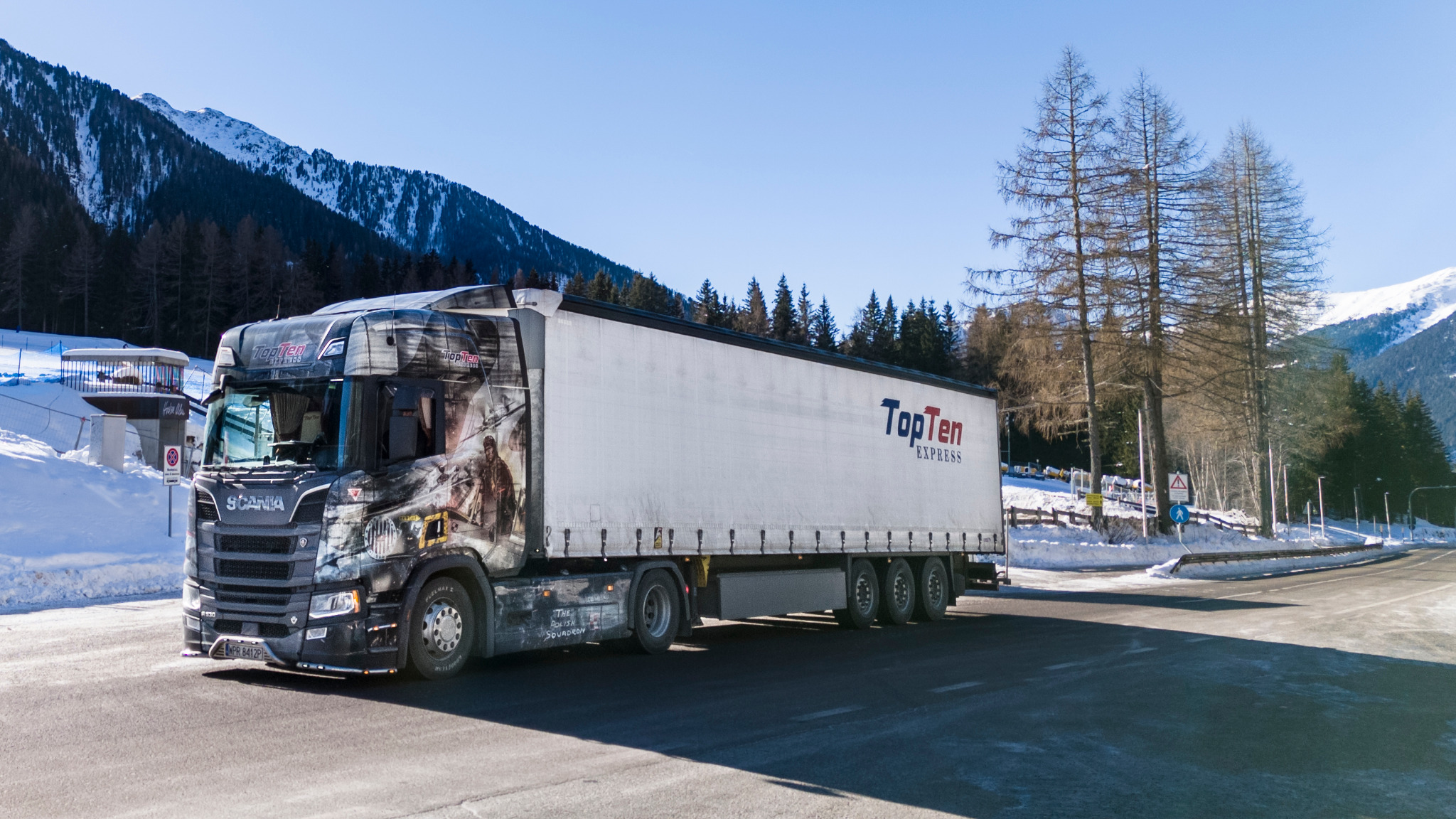 Czarno-biała ciężarówka Scania z grafiką 'TopTen Express' na tle ośnieżonych gór i drzew. Pojazd na drodze w słoneczny dzień.