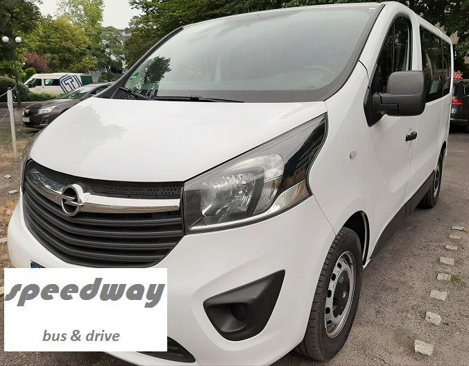 Biały Opel Vivaro z logo firmy transportowej na przednim planie, zaparkowany na brukowanym parkingu.