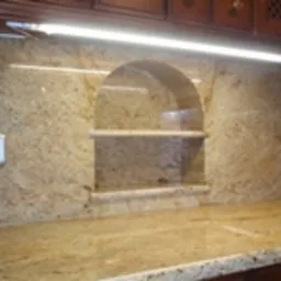 Blat i ściana wykończone granitem w odcieniach beżu i brązu, z wnęką z półkami o łukowatym kształcie. Widoczny włącznik elektryczny po lewej stronie i oświetlenie LED pod szafkami.