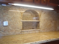 Blat i ściana wykończone granitem w odcieniach beżu i brązu, z wnęką z półkami o łukowatym kształcie. Widoczny włącznik elektryczny po lewej stronie i oświetlenie LED pod szafkami.