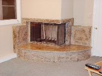 Wykończony kominek narożny z obudową z beżowego kamienia z wyraźną fakturą, drewniane drzwiczki kominkowe, widok z boku z fragmentem podłogi i pilota.