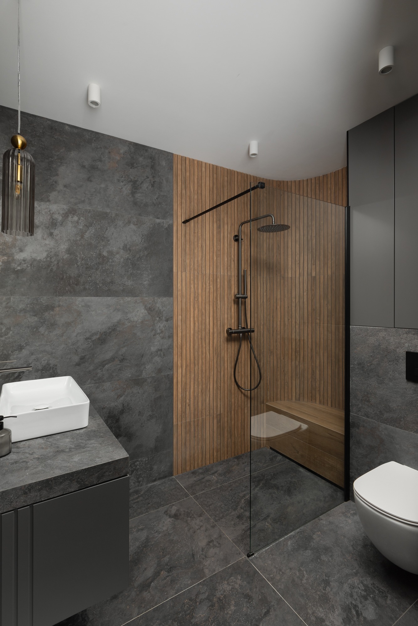 Nowoczesna łazienka z ciemnoszarą płytką imitującą kamień, drewnianą ścianą prysznicową z siedziskiem, białą umywalką nablatową i czarną armaturą.