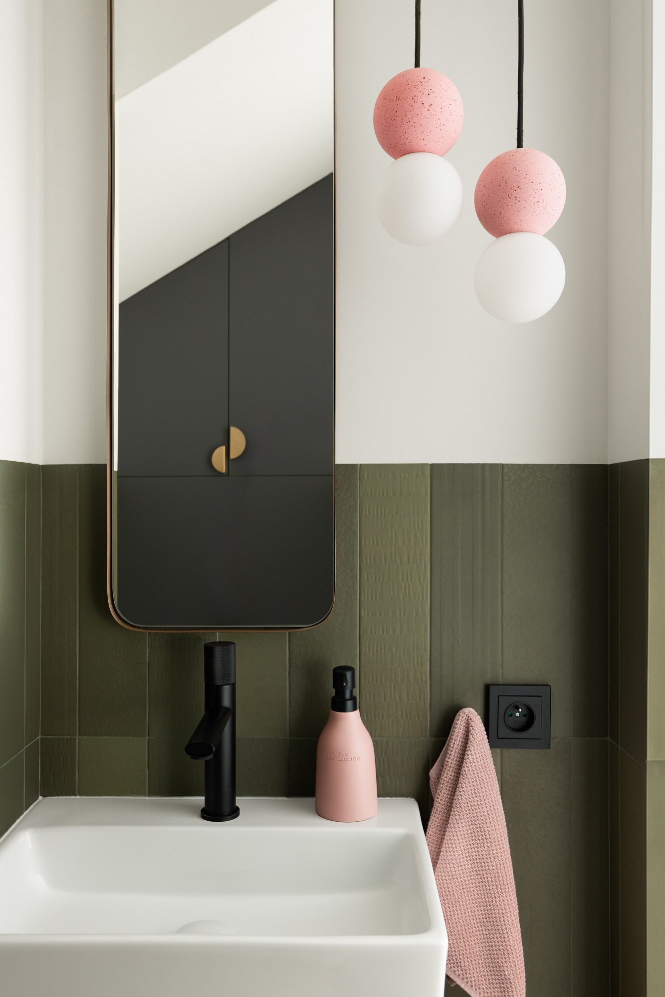 Nowoczesna łazienka z zielonymi, teksturowanymi płytkami, białym umywalką, czarną armaturą, różowym dozownikiem mydła i lampami sufitowymi z różowo-białymi kulami.