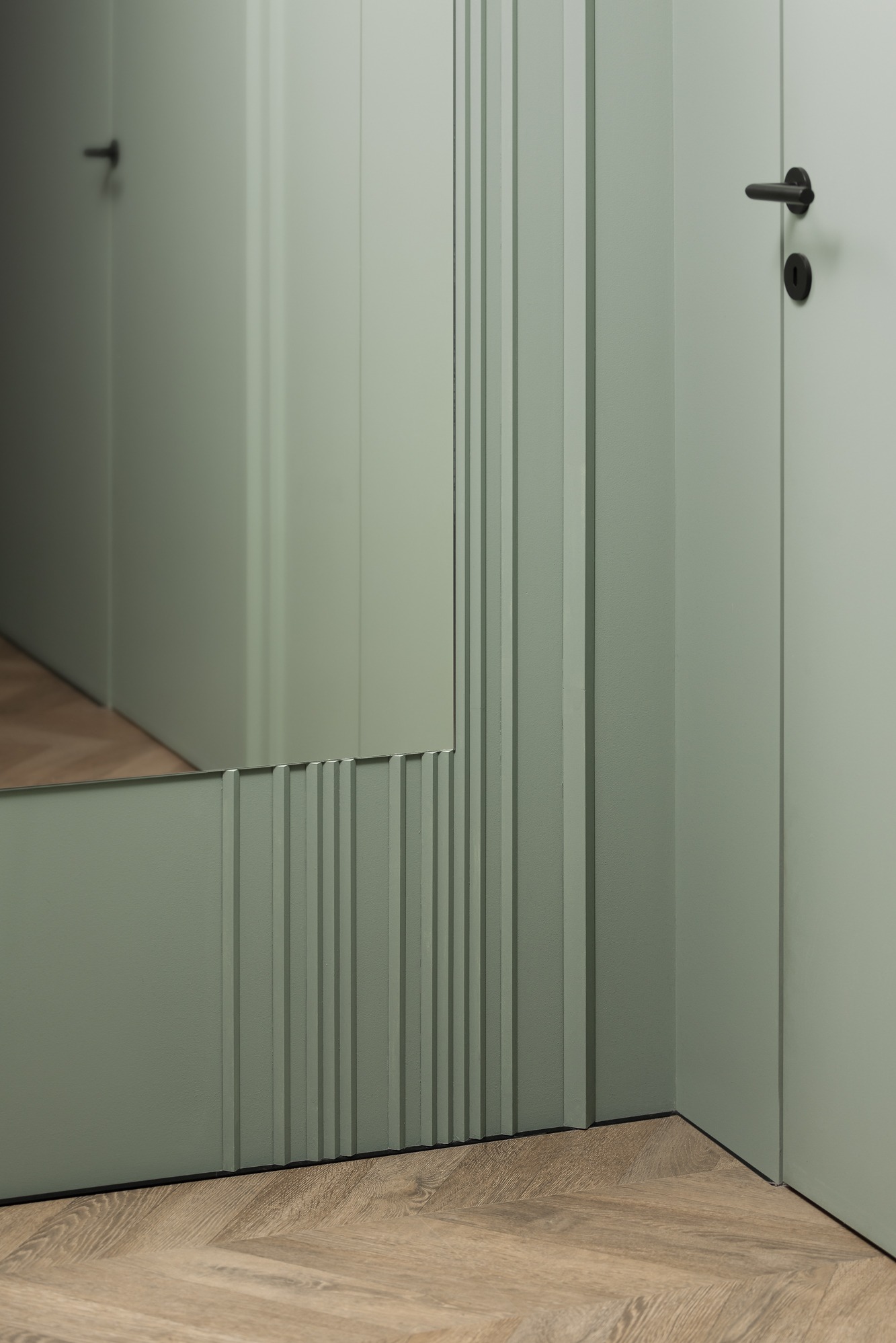 Fragment korytarza z lustrem z dekoracyjnymi pionowymi listwami w odcieniu stonowanej zieleni, parkiet ułożony we wzór jodełki.