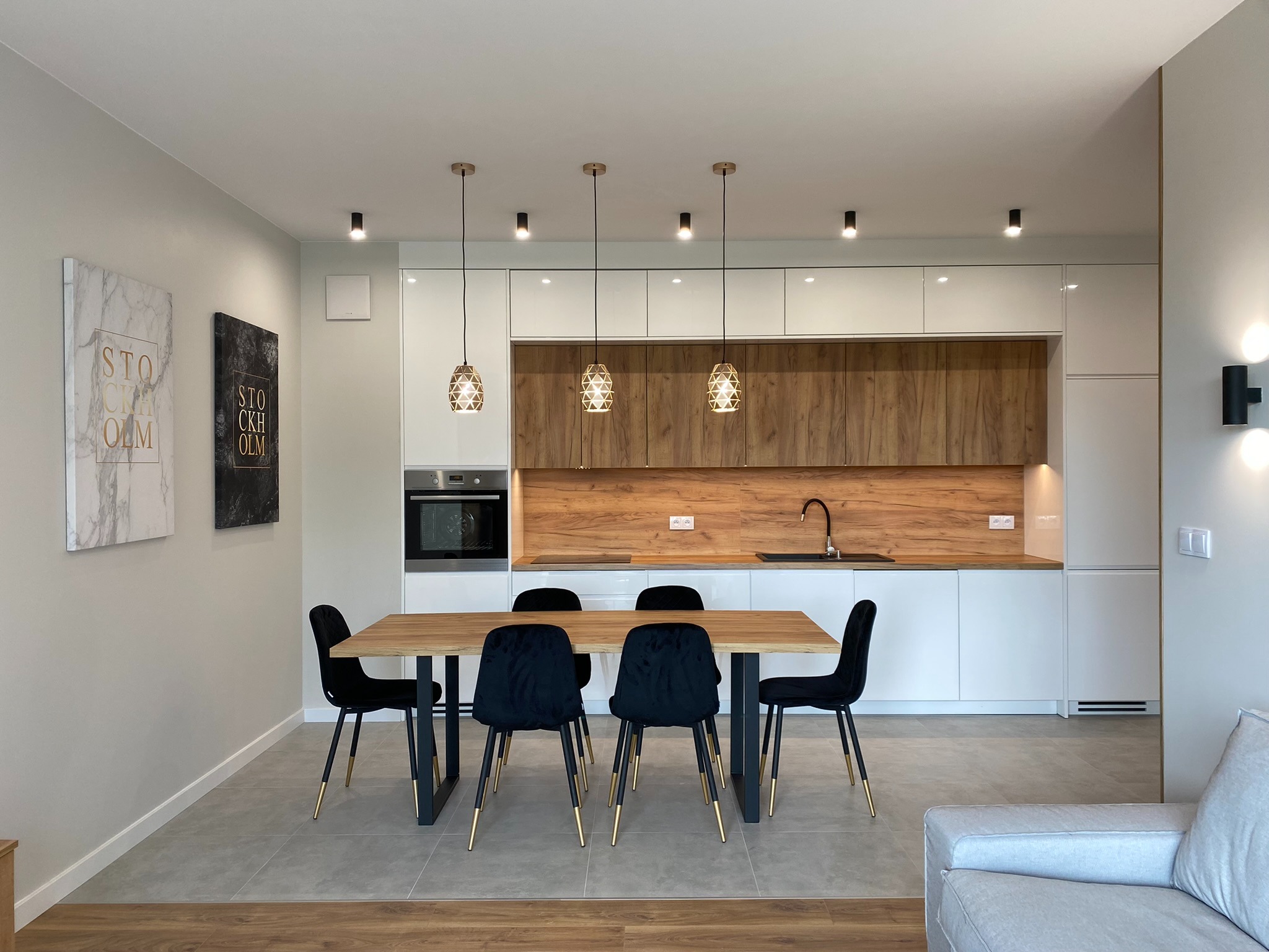Nowoczesna jadalnia z drewnianym stołem, czarnymi krzesłami z złotymi nogami i zabudową kuchenną z białymi frontami i drewnianymi akcentami, oświetlona wiszącymi lampami z ażurowymi kloszami.