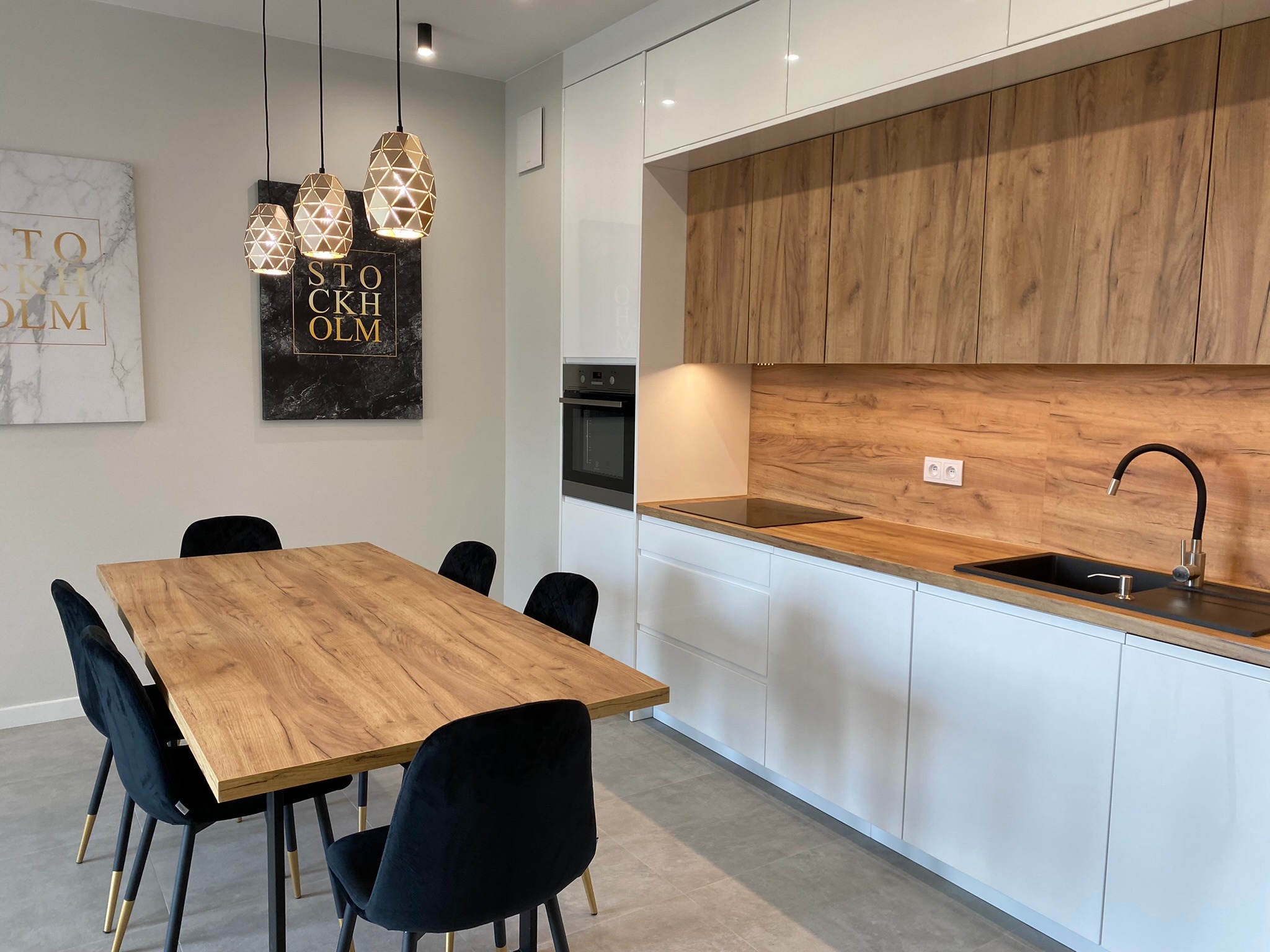 Nowoczesna kuchnia z drewnianymi i białymi frontami, drewnianym blatem, czarnym zlewem i baterią, oświetlona trzema lampami wiszącymi nad drewnianym stołem z czarnymi krzesłami.