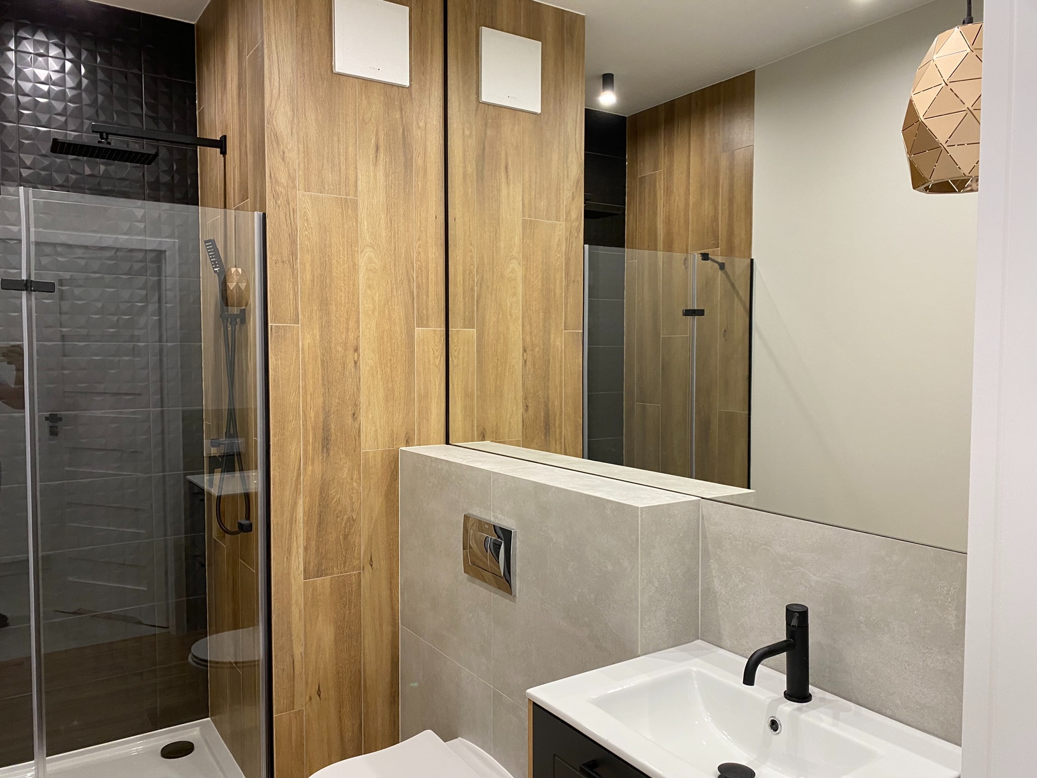 Nowoczesna łazienka z kabiną prysznicową typu walk-in, drewnopodobnymi płytkami na ścianach, czarną armaturą i geometryczną lampą wiszącą.