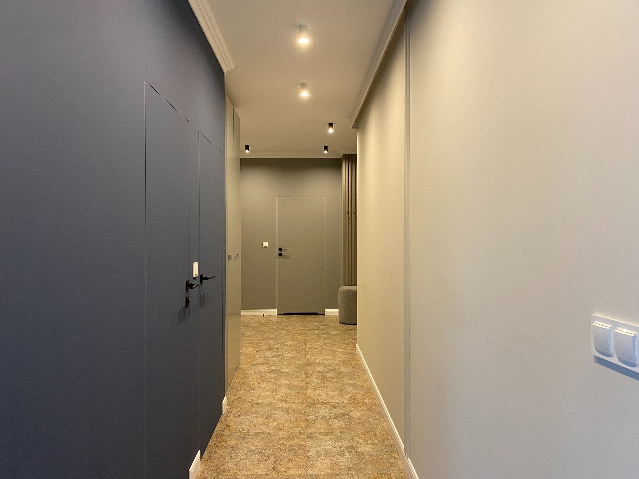 Wnętrze korytarza w odcieniach szarości i beżu, z ciemnogranatową szafą w zabudowie, punktowym oświetleniem sufitowym i podłogą imitującą kamień.