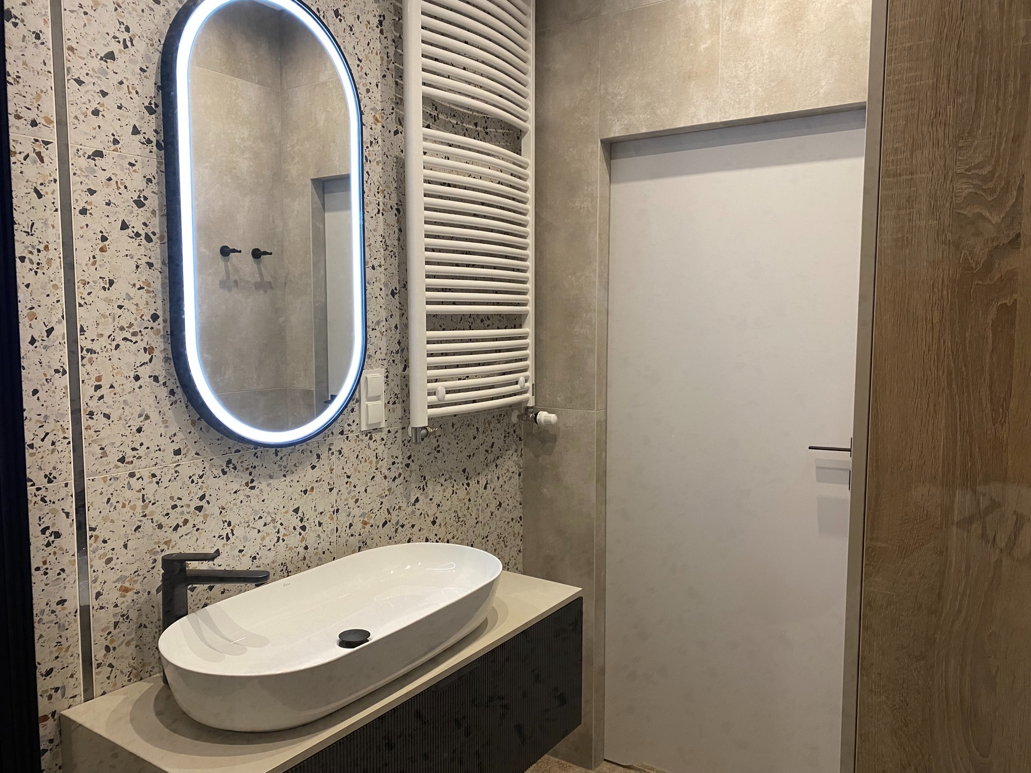 Nowoczesna łazienka z owalną umywalką nablatową, czarną armaturą, podświetlanym lustrem i grzejnikiem drabinkowym na ścianie z mozaiką.