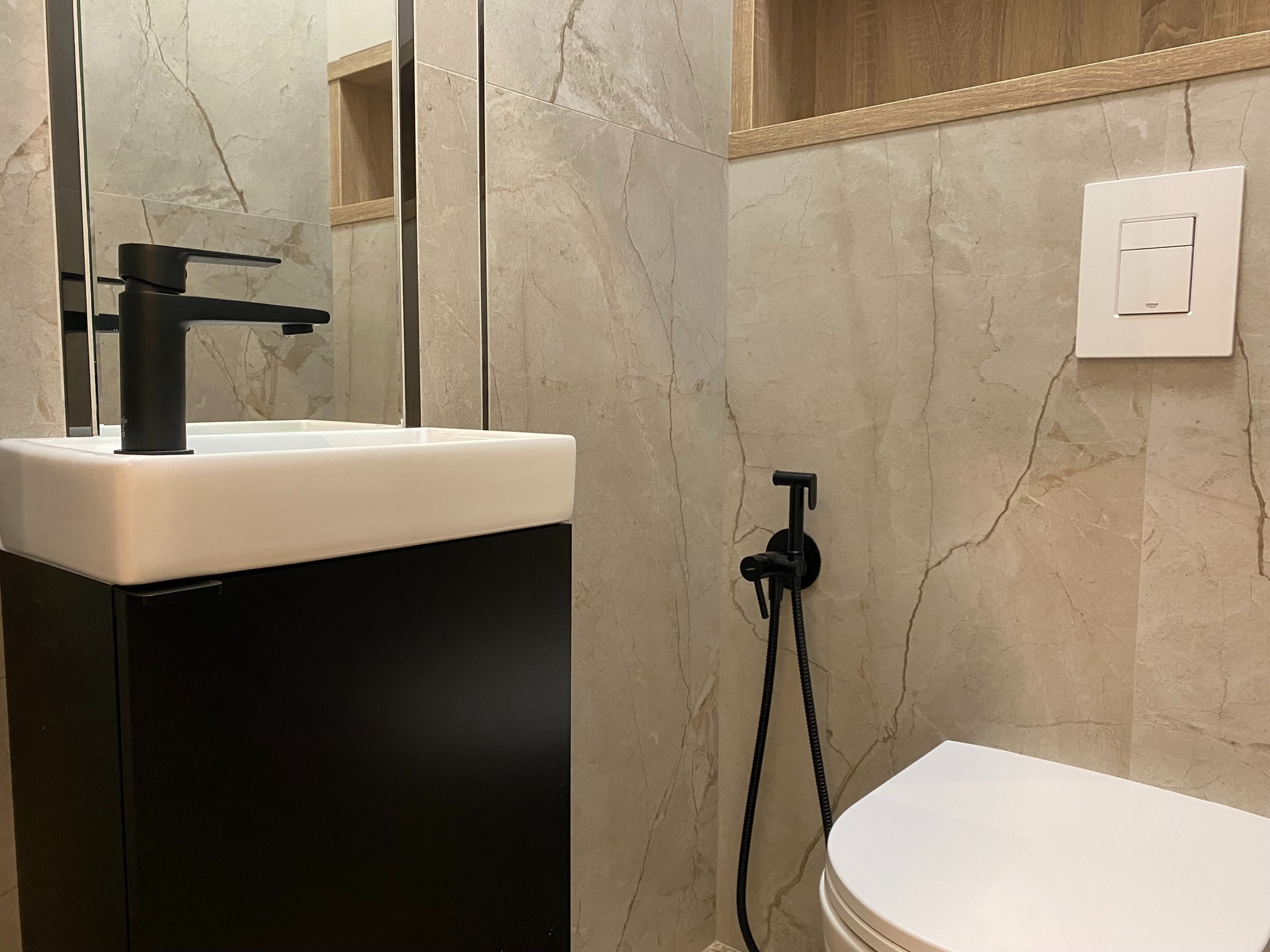 Umywalka z czarną armaturą, bidetka i toaleta w łazience wykończonej płytkami imitującymi marmur z widocznym w tle lustrem i półką.
