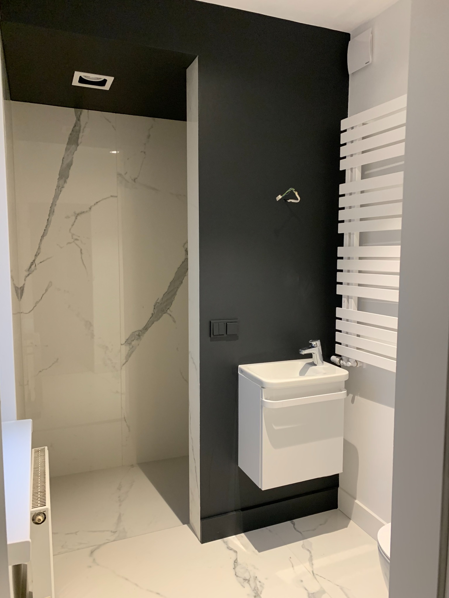 Nowoczesna łazienka z czarną ścianą, białą umywalką wiszącą i grzejnikiem drabinkowym, wykończona płytkami imitującymi marmur.