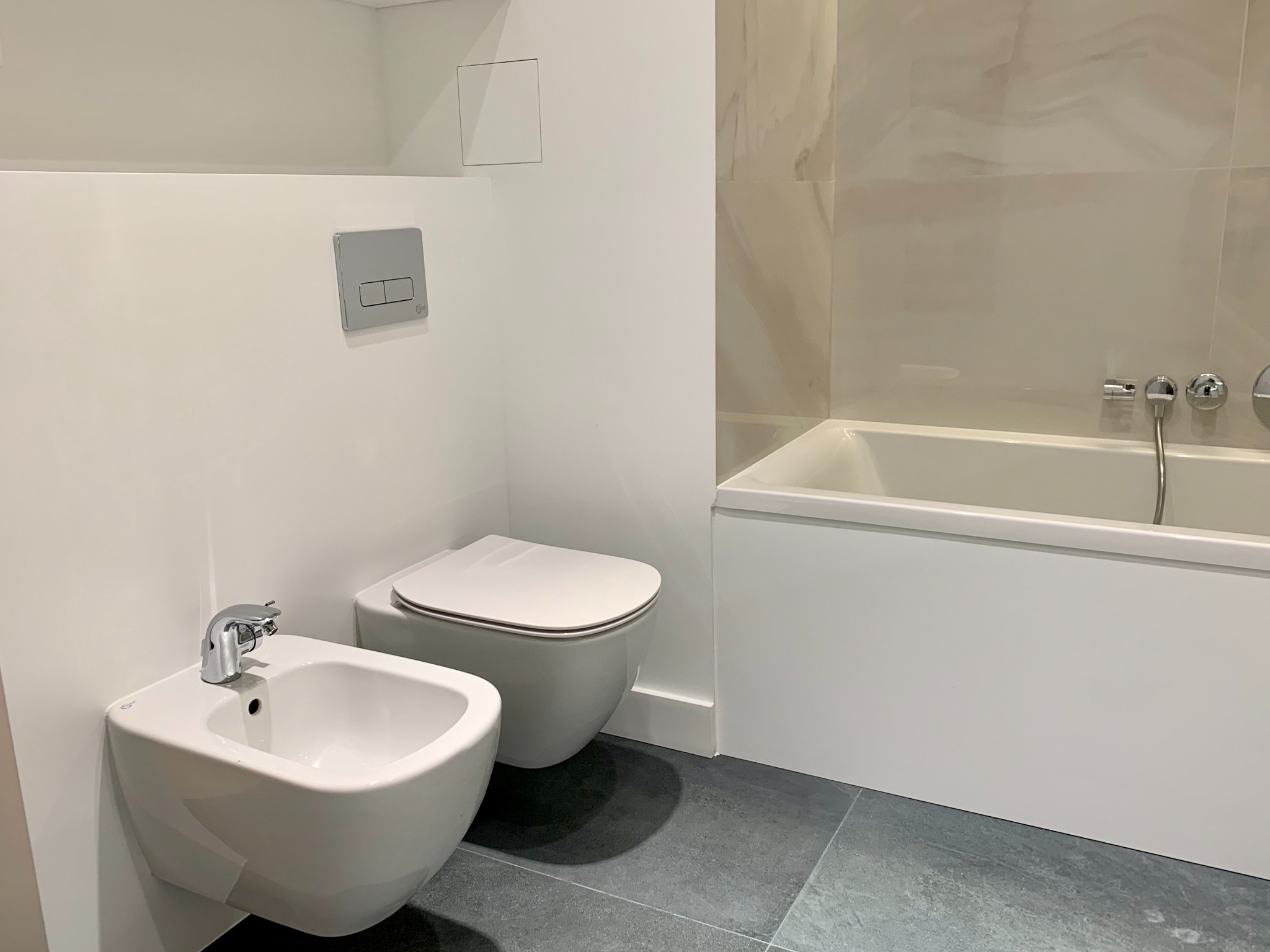 Nowoczesna łazienka z białą ceramiką sanitarną: wisząca toaleta i bidet z chromowaną armaturą, obok zabudowana wanna z marmurowymi płytkami na ścianie.