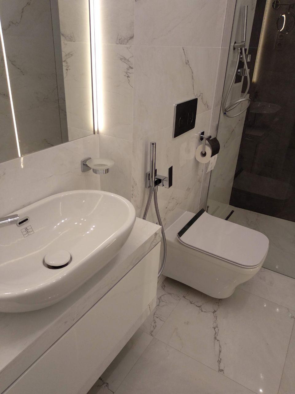 Nowoczesna łazienka wykończona białym marmurem, owalna umywalka nablatowa, podwieszana toaleta z czarnym akcentem i kabina prysznicowa typu walk-in.