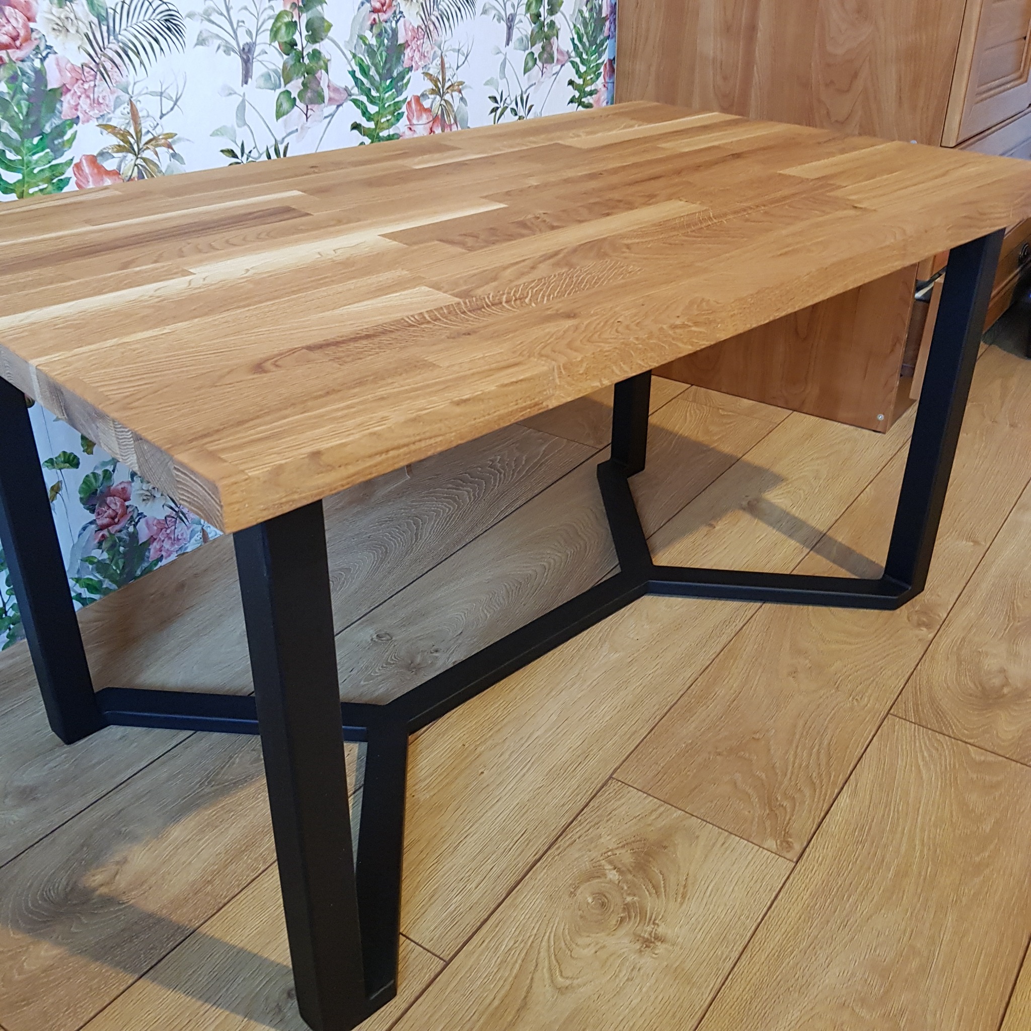 Stół z drewnianym blatem i czarnymi, metalowymi nogami o geometrycznym kształcie, wykonany na zamówienie w Krakowie. W tle widoczna ściana z kwiatowym wzorem.