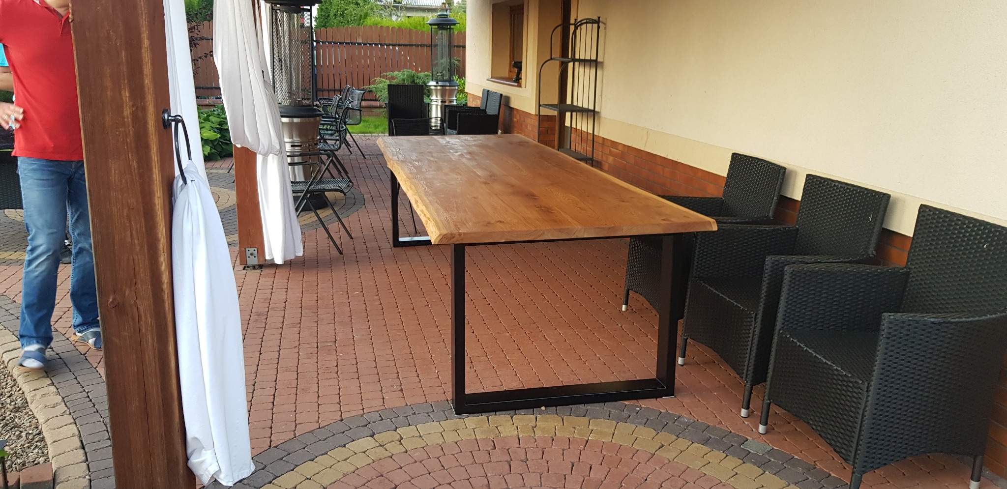 Rustykalny stół z blatem z litego drewna i czarnymi metalowymi nogami, ustawiony na tarasie z ceglaną kostką, otoczony czarnymi krzesłami z technorattanu i białymi zasłonami.