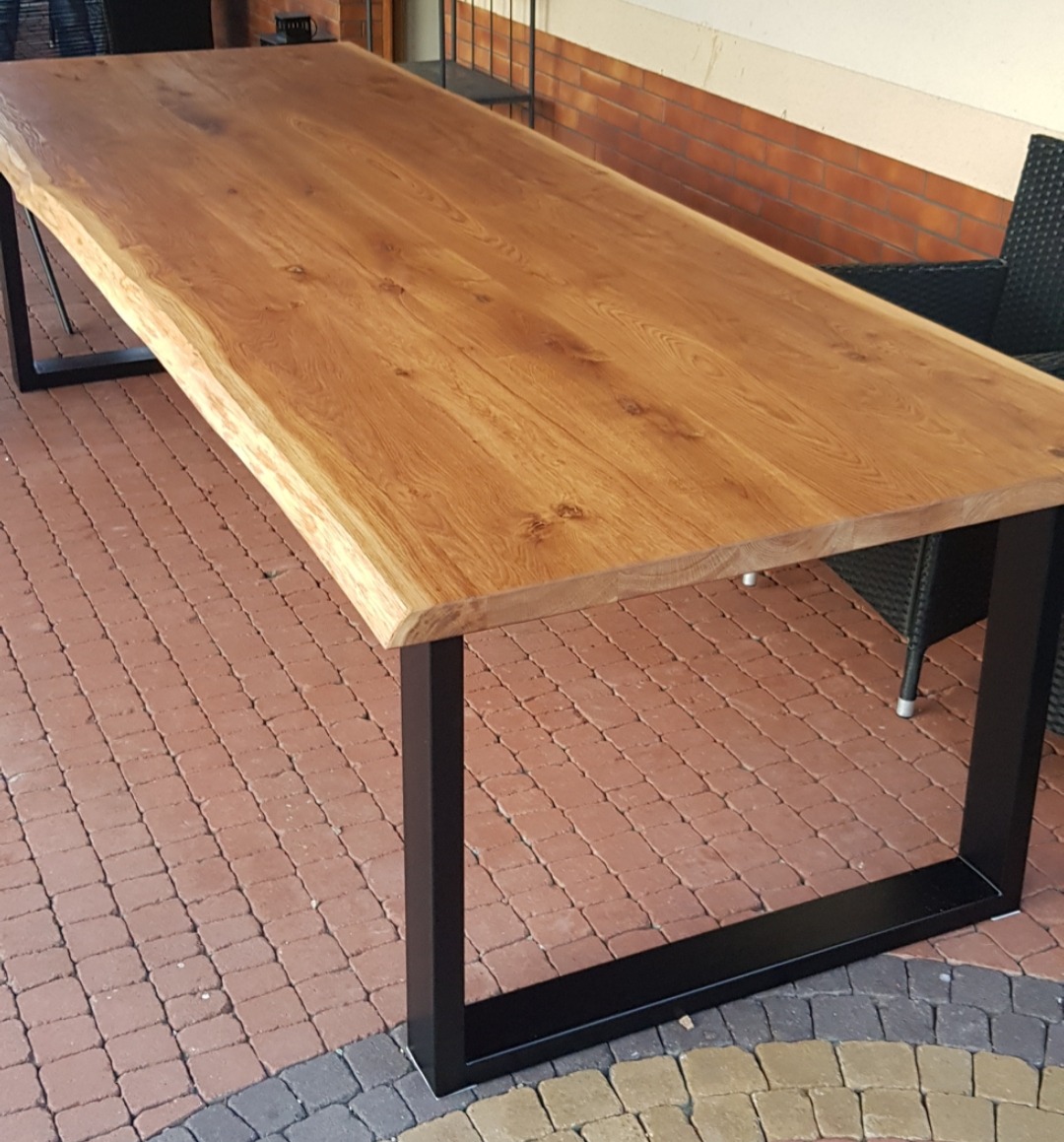 Stół z blatem z litego drewna dębowego o nieregularnej krawędzi, wsparty na czarnych, metalowych nogach o kwadratowym przekroju, ustawiony na ceglanym patio.