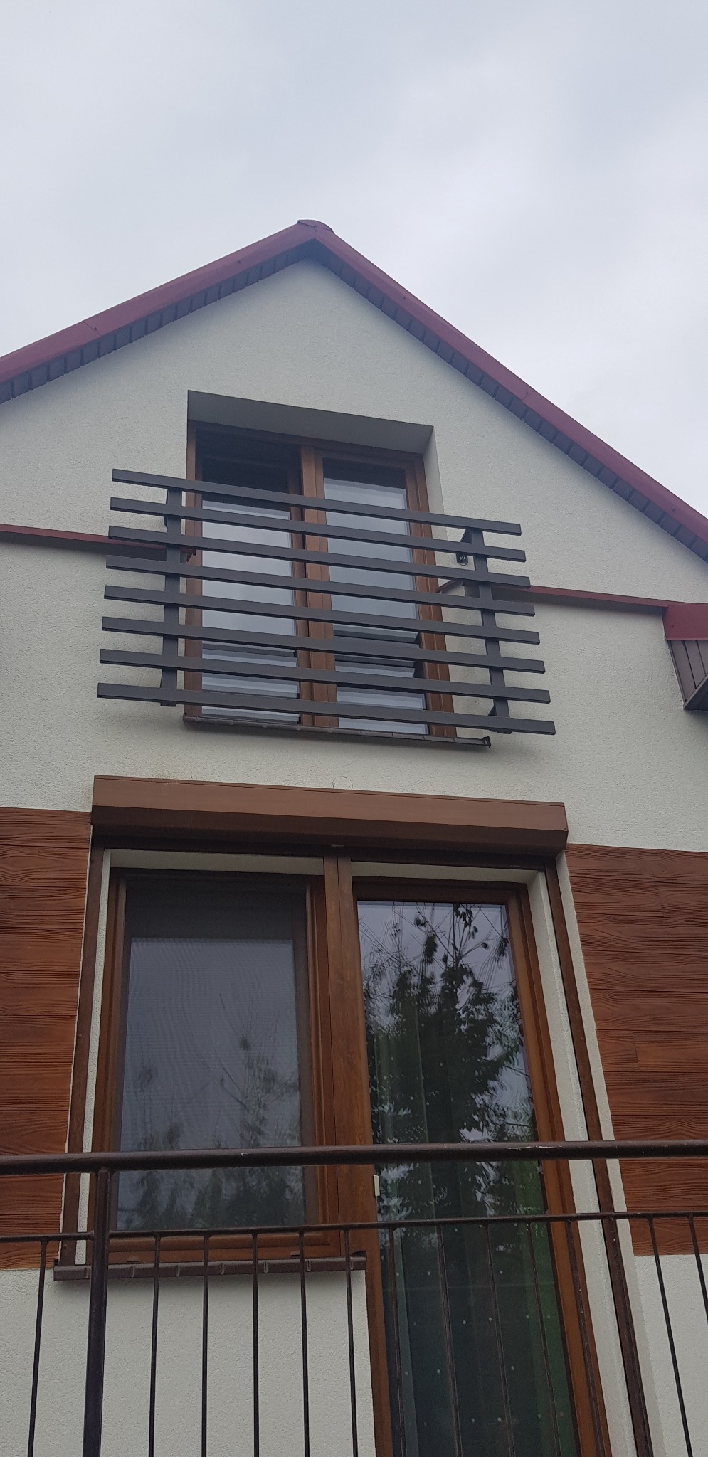 Fasada domu z nowoczesną, poziomą balustradą balkonową w kolorze grafitowym, widoczna stolarka okienna w kolorze drewna i okładzina elewacyjna imitująca drewno.