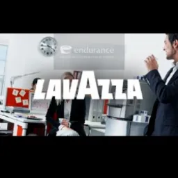 Biuro z pracownikami, ekspres do kawy w tle, logo Lavazza na pierwszym planie, zegar na ścianie i logo firmy Endurance. Mężczyzna w garniturze pije kawę z białego kubka.