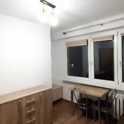 Jasne wnętrze pokoju po odświeżeniu, z białymi ścianami, drewnianymi meblami i oknami z roletami, widoczne ślady świeżo wykonanych prac wykończeniowych.