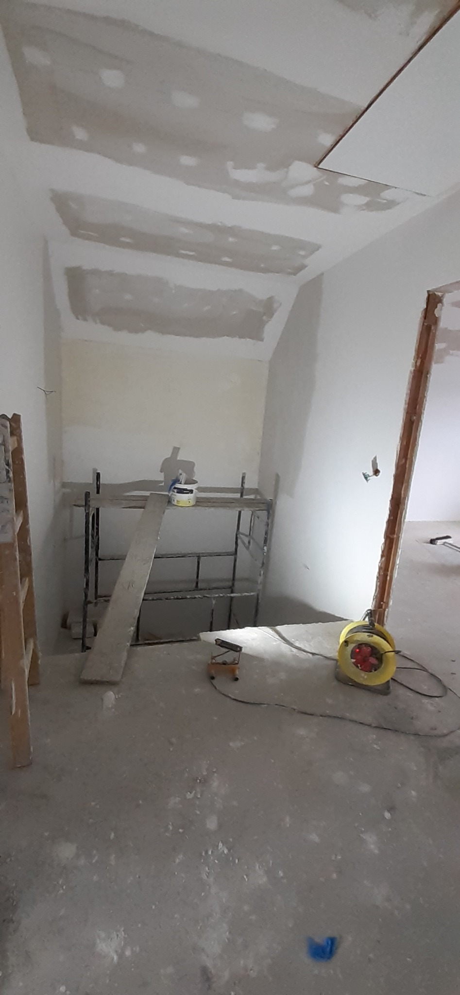 Wnętrze w trakcie prac wykończeniowych: ściany i sufit z widocznymi łatami, rusztowanie z wiadrem i narzędziami, przedłużacz elektryczny na podłodze.