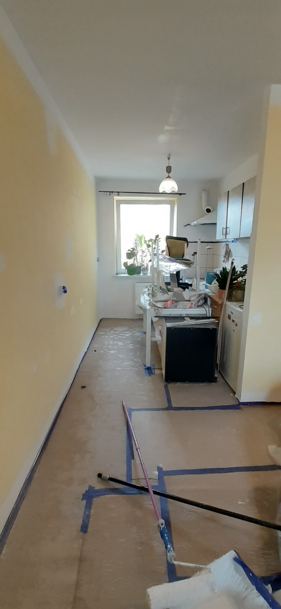 Przygotowania do odświeżenia wnętrza: pokój z zabezpieczoną taśmą podłogą, częściowo pomalowanymi ścianami w odcieniach żółci, widoczne wałki i kije malarskie, w tle okno z roślinami i fragment...