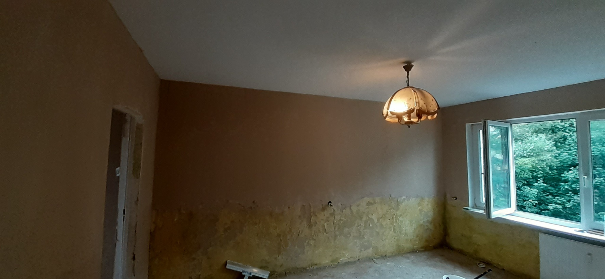 Pokój w trakcie remontu z widocznymi śladami po usuniętej starej warstwie, przygotowany do dalszych prac wykończeniowych, z otwartym oknem i wiszącą lampą w stylu retro.