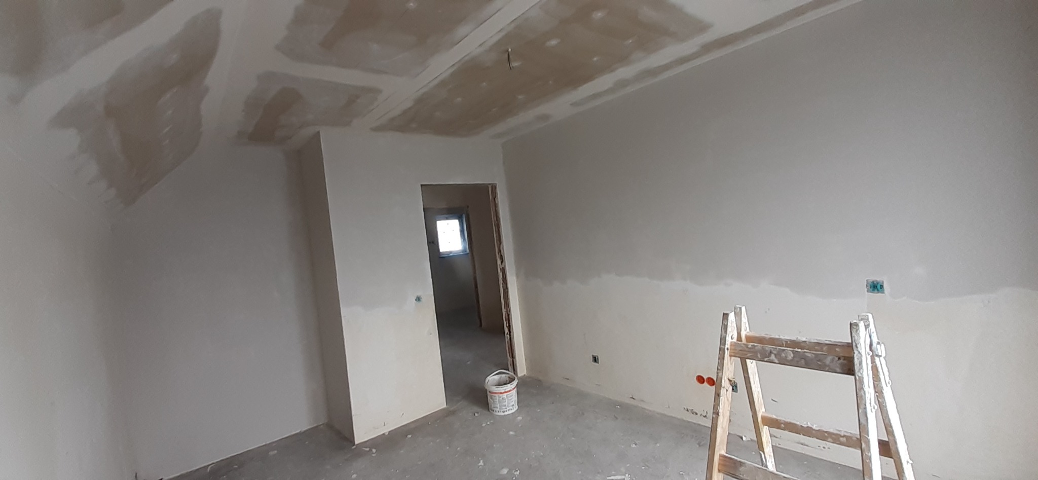 Pokój w trakcie remontu: ściany i sufit po nałożeniu gładzi, widoczna drabina malarska, wiadro z farbą i niedokończone wykończenie wokół gniazdka elektrycznego.