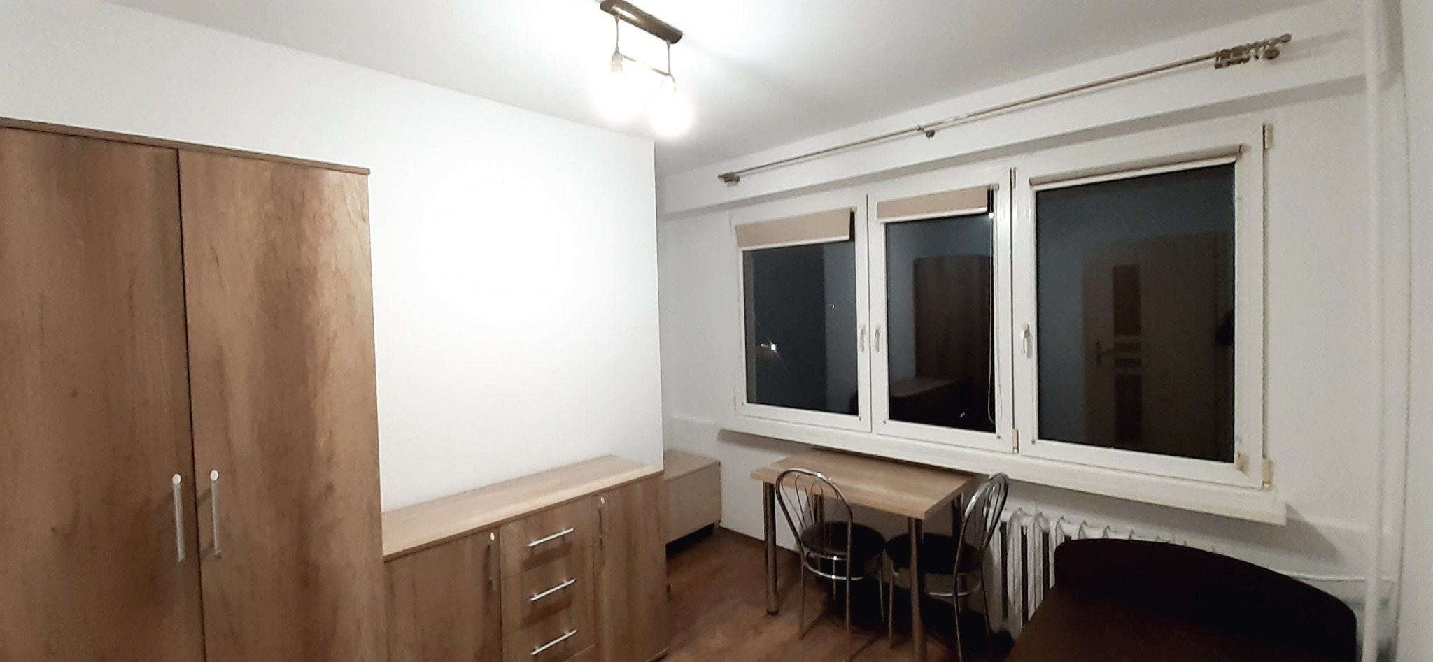 Jasne wnętrze pokoju po odświeżeniu, z białymi ścianami, drewnianymi meblami i oknami z roletami, widoczne ślady świeżo wykonanych prac wykończeniowych.