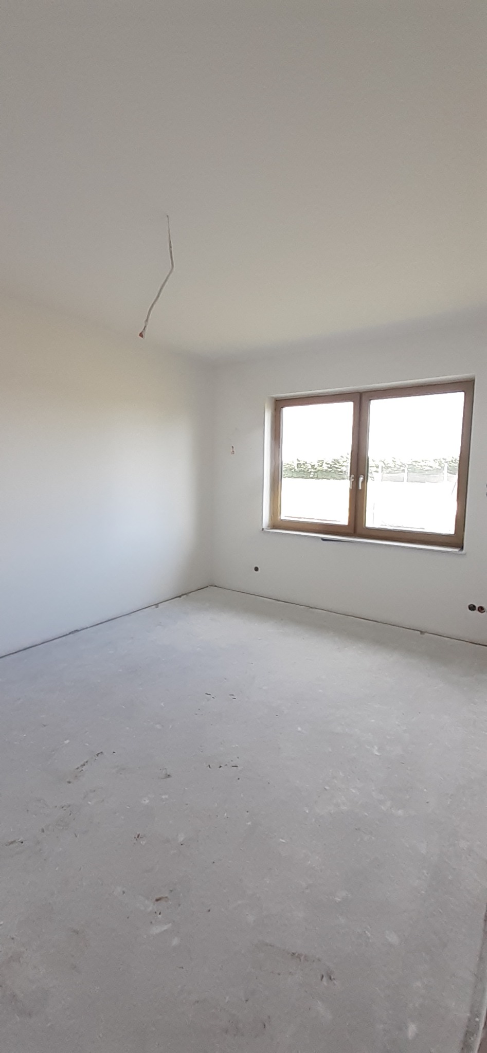 Pusty pokój po remoncie z surowymi ścianami, betonową podłogą i niedokończonymi instalacjami elektrycznymi, widok na okno z drewnianą ramą.