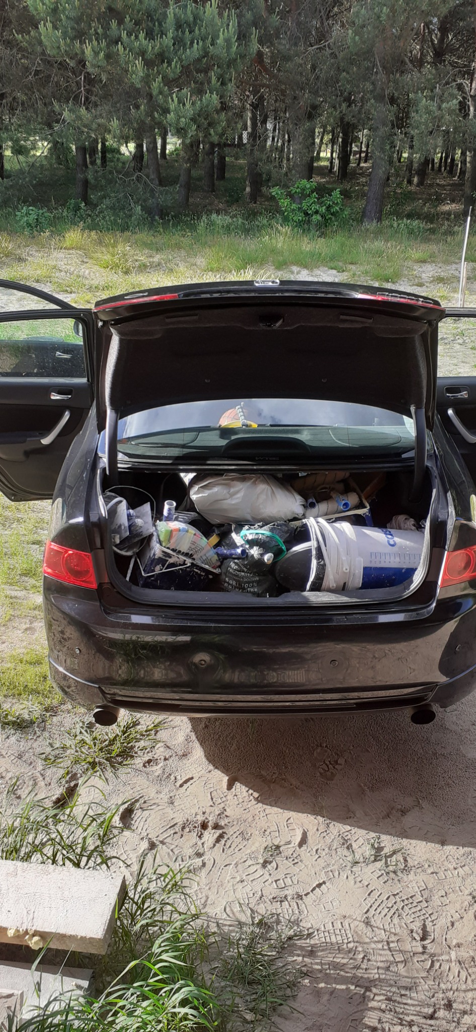 Otwarty bagażnik czarnego samochodu osobowego zapakowany materiałami i narzędziami budowlanymi, widok z tyłu na tle lasu.