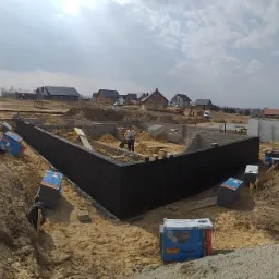 Wykop pod fundamenty domu jednorodzinnego z częściowo wylanymi ścianami, zabezpieczonymi czarną izolacją. Na budowie widoczny pracownik z taczką i porozrzucane kartony z materiałami izolacyjnymi.