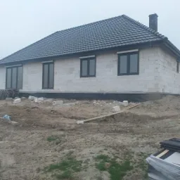 Niedokończony parterowy dom z białego pustaka z ciemną dachówką, czarnymi oknami i kominem, widoczne fundamenty i teren budowy.