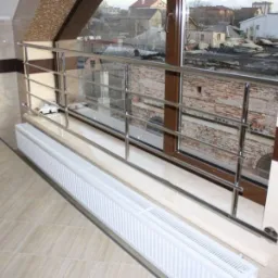 Barierka ze stali nierdzewnej zamontowana przy oknie na poddaszu, widok na zabudowania przez okno, grzejnik pod oknem.