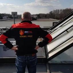 Mężczyzna w kurtce z logo firmy serwisującej systemy bezpieczeństwa pożarowego, stojący na dachu obok otwartego świetlika dachowego.