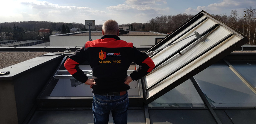 Mężczyzna w kurtce z logo firmy serwisującej systemy bezpieczeństwa pożarowego, stojący na dachu obok otwartego świetlika dachowego.