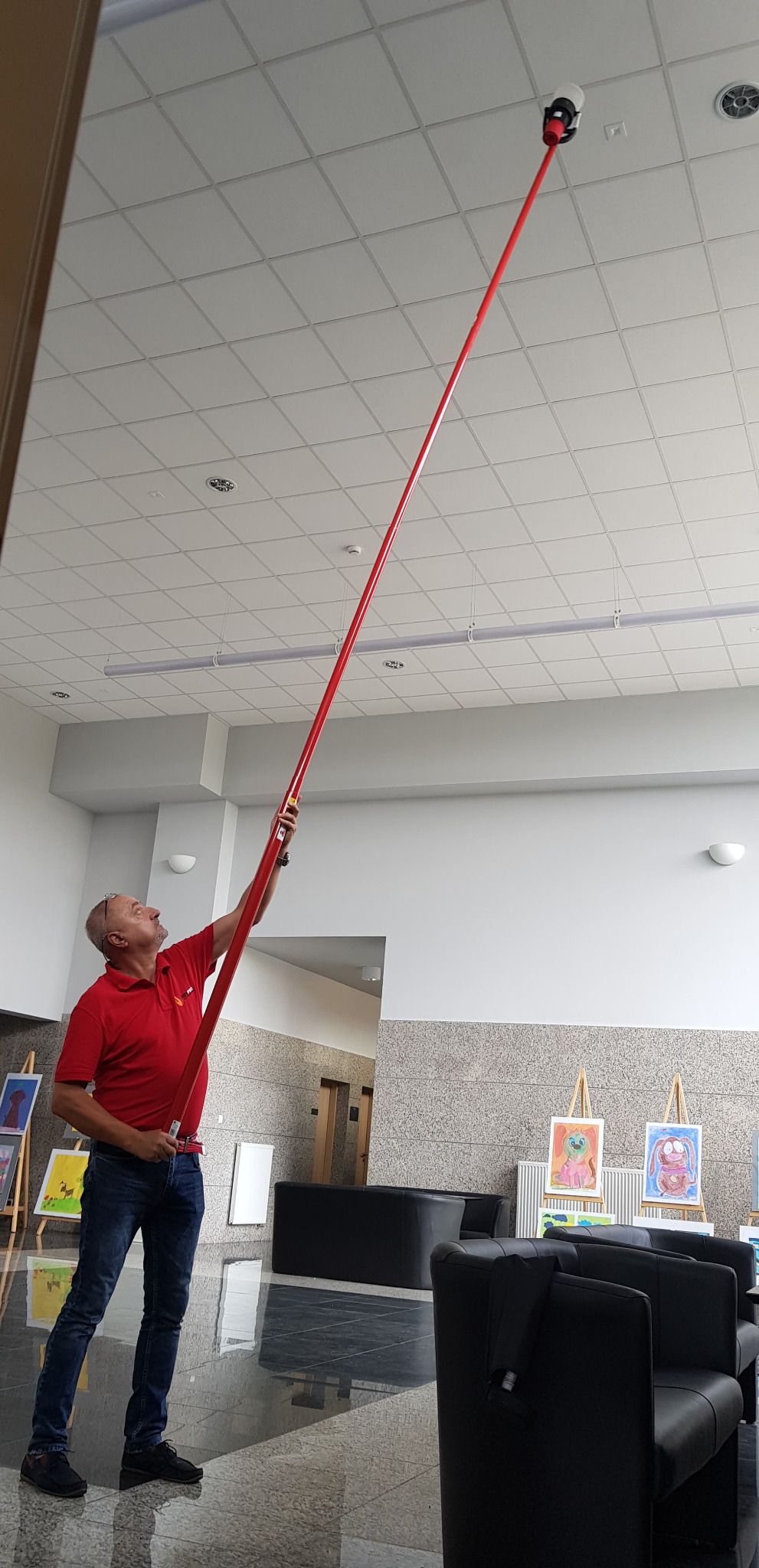 Mężczyzna w czerwonej koszulce sprawdza czujnik dymu na suficie za pomocą długiego, czerwonego drążka teleskopowego.