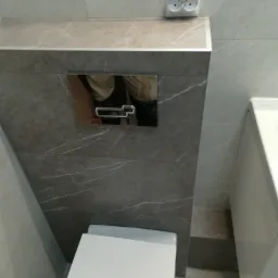 Szary, marmurkowy stelaż podtynkowy WC z chromowanym przyciskiem spłukującym, biała misa WC i gniazdko elektryczne w wykończonej łazience.