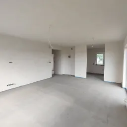 Wnętrze domu po tynkowaniu maszynowym, widoczne gładkie, jasnoszare ściany, surowy beton na podłodze, wystające kable elektryczne i okno w głębi