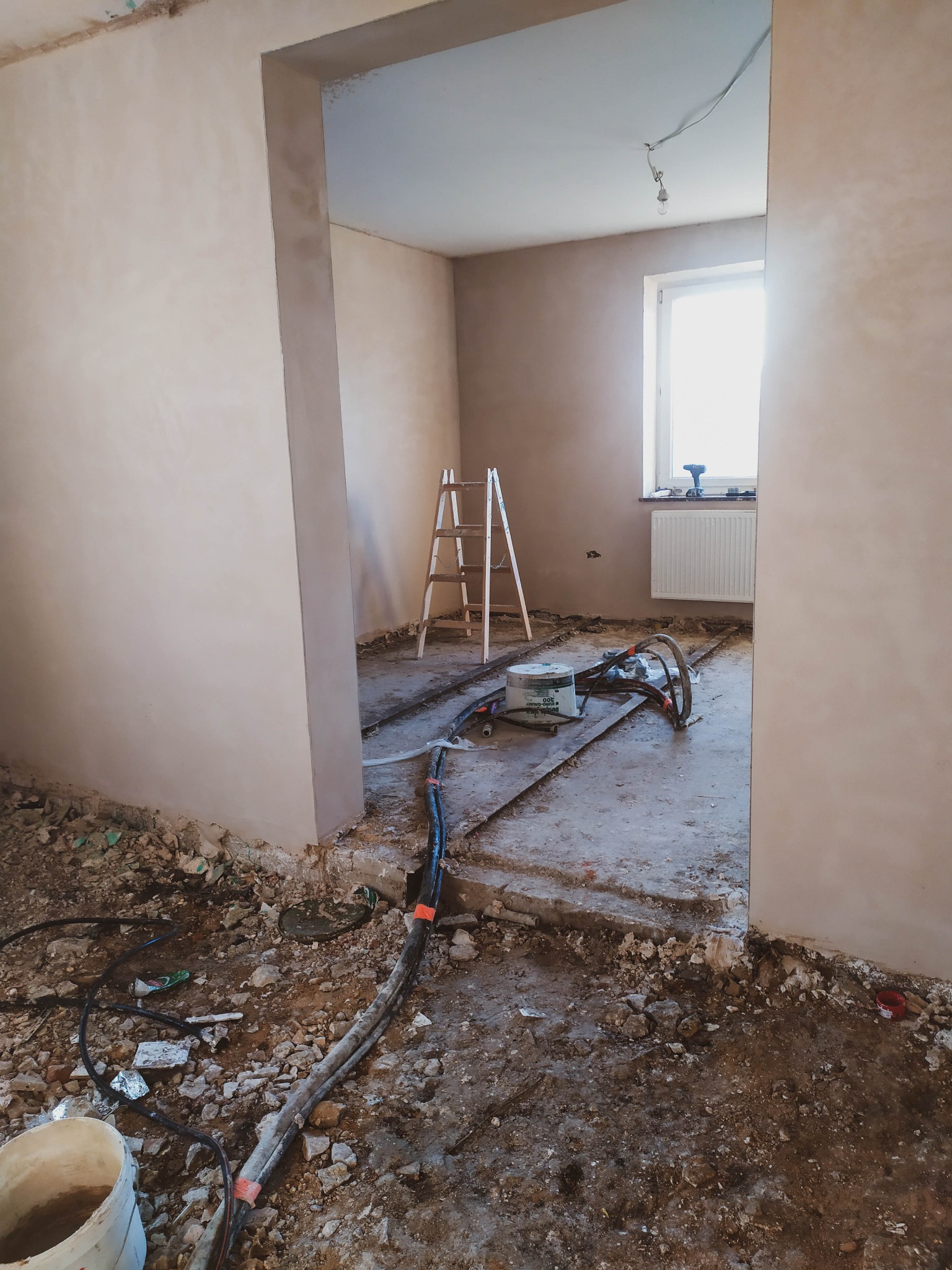 Wnętrze w trakcie prac wykończeniowych: świeżo otynkowane ściany, drabina oparta o ścianę, przewody i maszyna leżące na podłodze w pokoju z oknem.