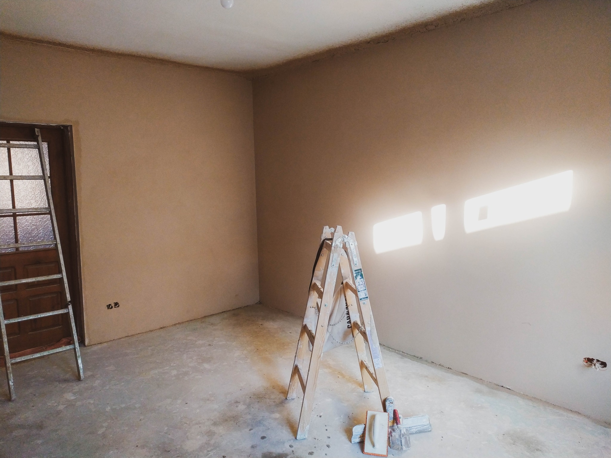 Pokój po nałożeniu tynku maszynowego, widoczne ślady po gniazdkach, drabina oparta o ścianę i narzędzia murarskie na podłodze, ślady po tynku na podłodze.