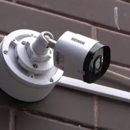 Biała kamera monitoringu marki Kenik zamontowana na brązowej ceglanej ścianie, z widocznymi elementami mocującymi i kablem w białej listwie.