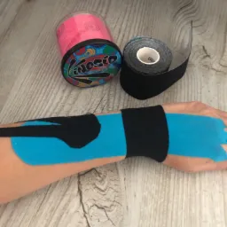 Ręka z aplikacją taśm kinezjologicznych w kolorze niebieskim i czarnym, obok dwie rolki taśm na drewnianym tle.