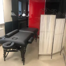 Pusta, czarna leżanka z regulowanym zagłówkiem i podłokietnikami, ustawiona w gabinecie z beżowymi ścianami i dekoracyjnym parawanem w tle.