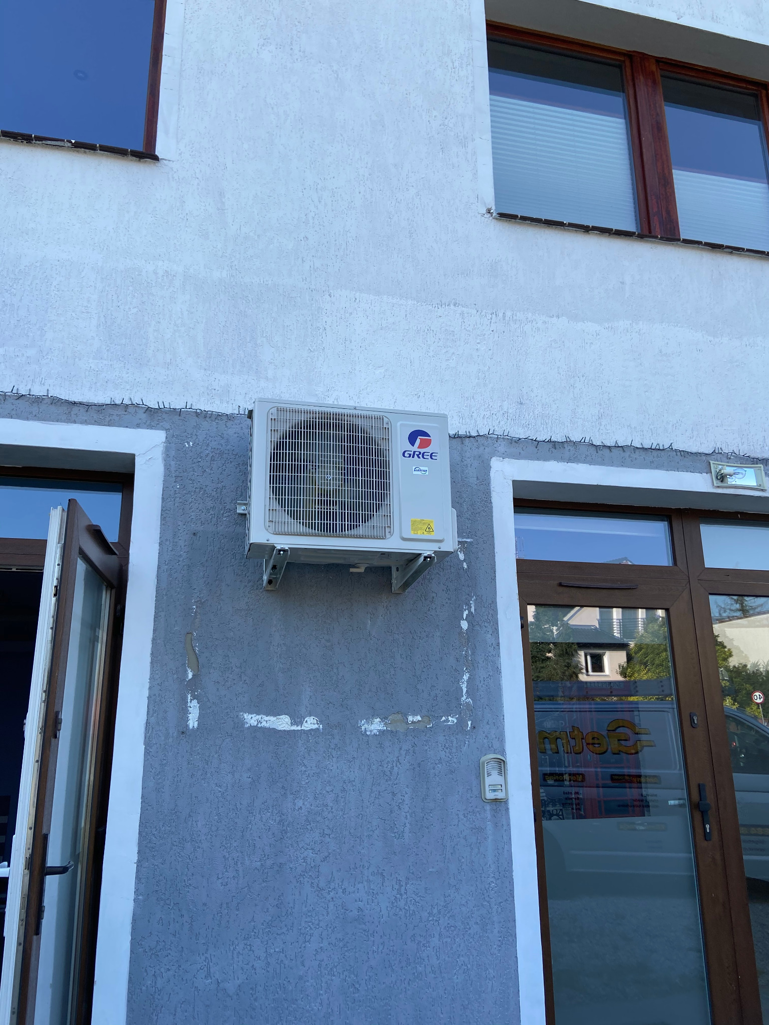 Biała jednostka zewnętrzna klimatyzatora marki Gree zamontowana na szarej ścianie budynku z widocznymi śladami po poprzednich instalacjach, tuż obok otwartych drzwi wejściowych.