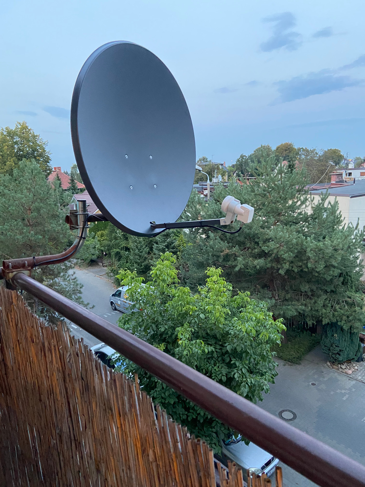 Szara antena satelitarna zamontowana na balkonie z widokiem na ulicę i zieleń, z metalowymi elementami mocującymi i brązową balustradą.