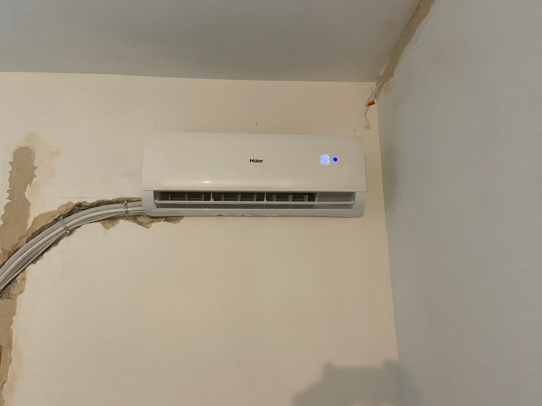 Biały klimatyzator Haier zamontowany na ścianie z widocznymi śladami po instalacji, odsłonięte przewody elektryczne przymocowane klamrami do ściany, niedokończone wykończenie ściany w miejscu montażu.