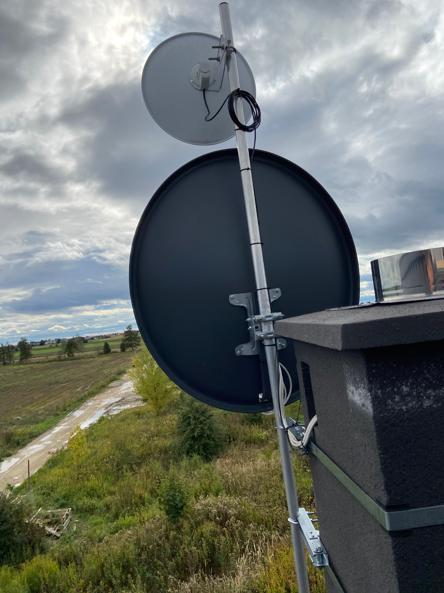 Dwie anteny satelitarne zamontowane na metalowym maszcie, widoczne przewody, tło to pochmurne niebo i krajobraz wiejski z polami i lasami.